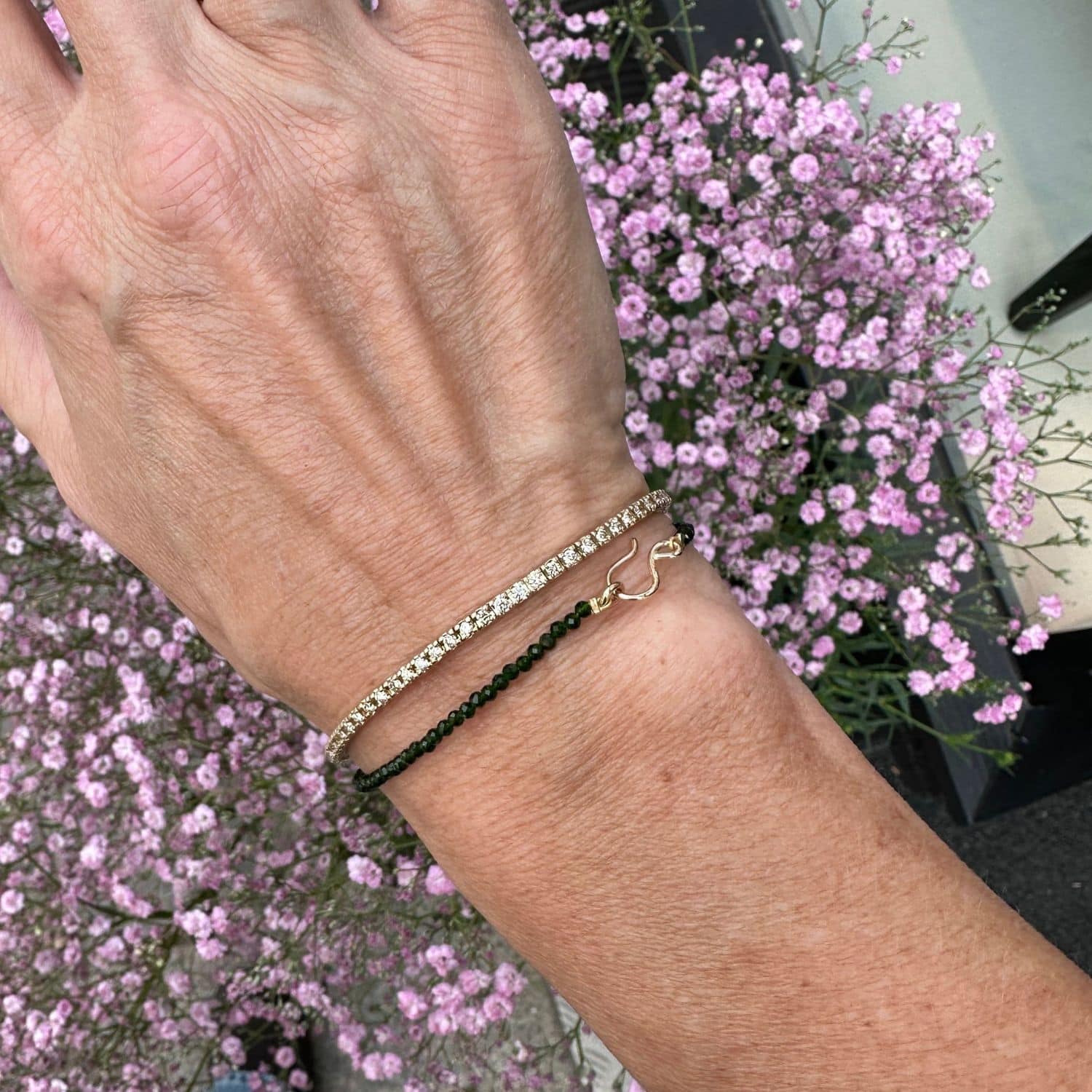 Armbånd med grøn agat og 14 kt guld S-lås, båret på en hånd med baggrund af lyserøde blomster.