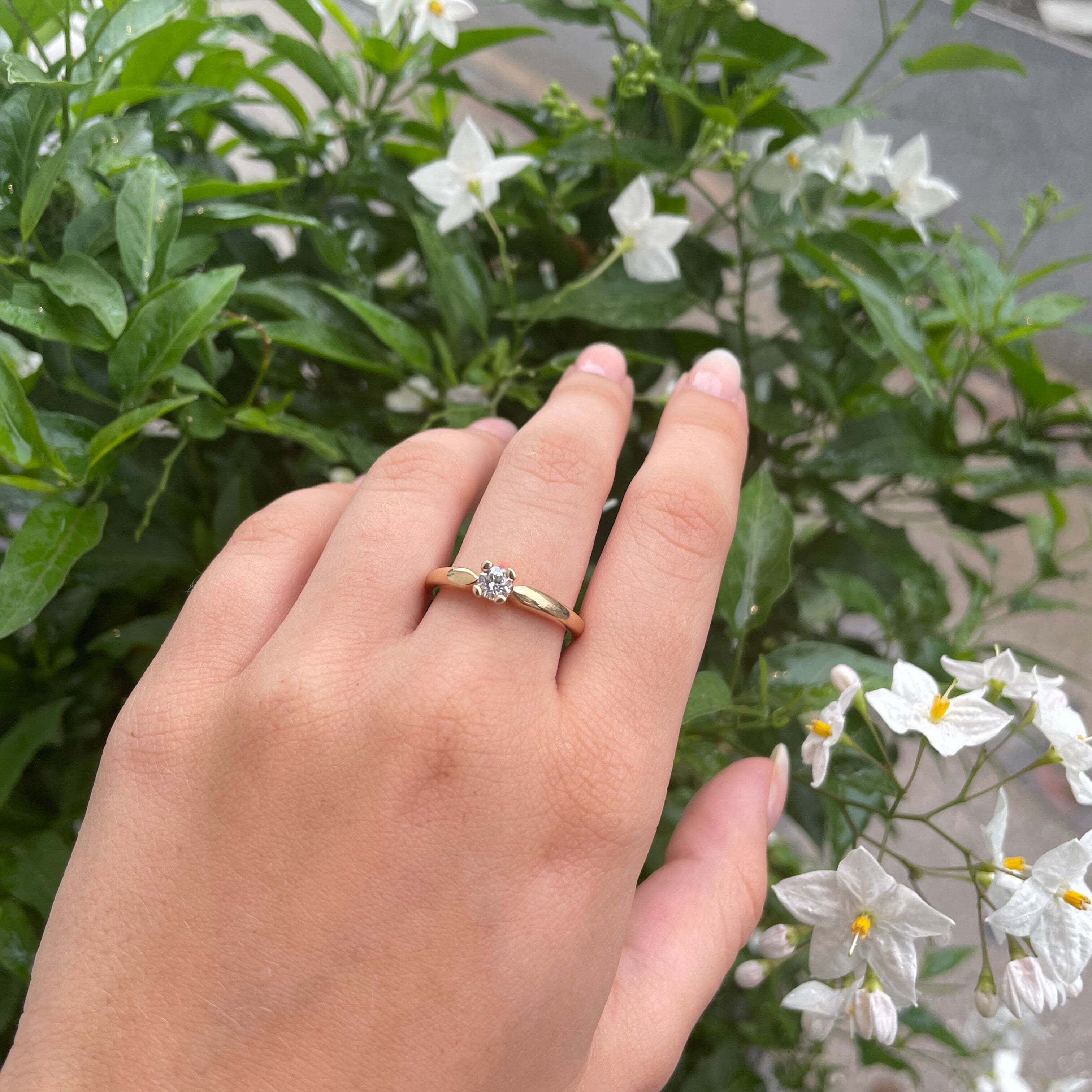 Forlovelsesring i guld med en brillant på en hånd, omgivet af hvide blomster i baggrunden.